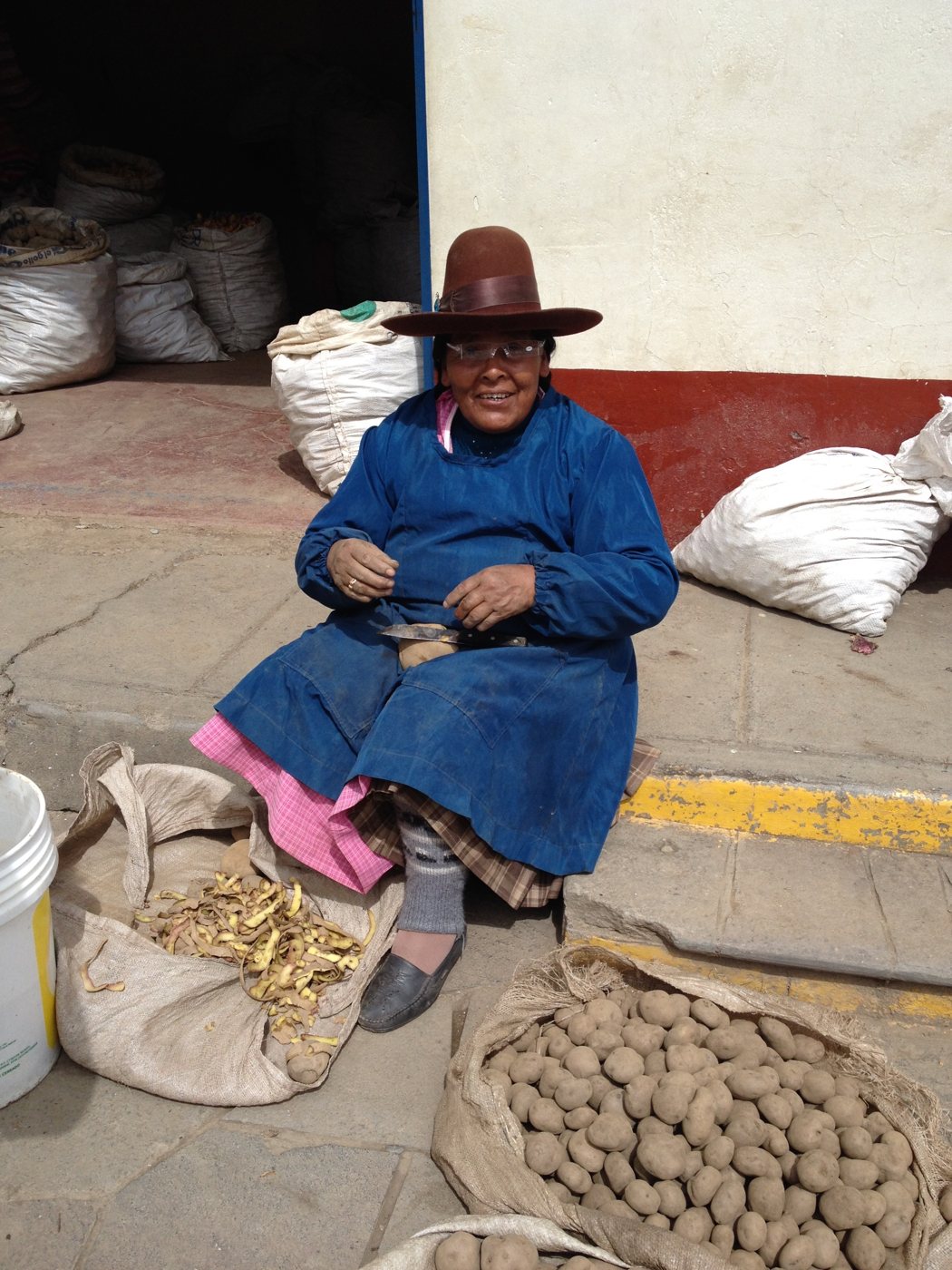 A woman in a brown felt hat and blue apron smiles in her new reading glasses while peeling potatoes on a market sidewalk